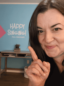 a picture of Nikki smiling and pointing to a decal on the wall behind her that says Happy Singing